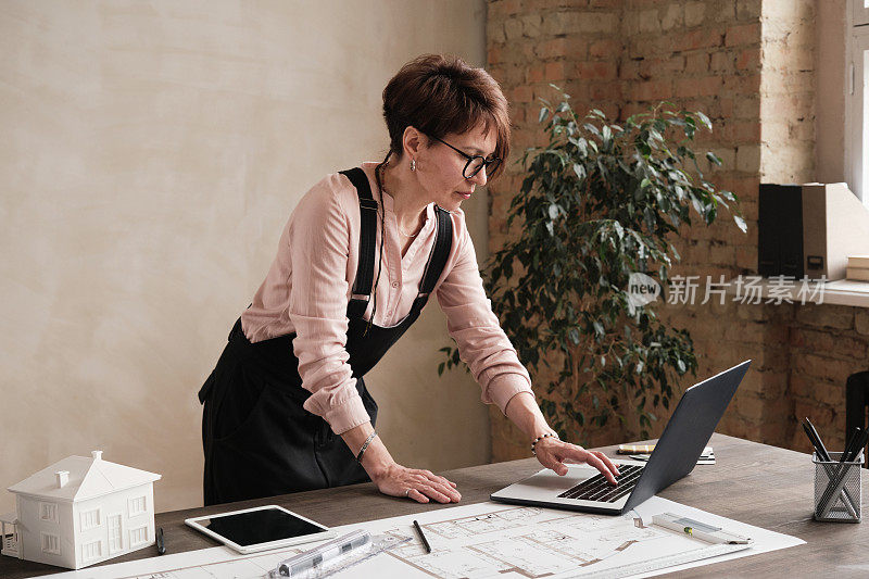 专注的女性建筑设计师戴着眼镜站在办公桌前，用笔记本电脑编辑蓝图和文件