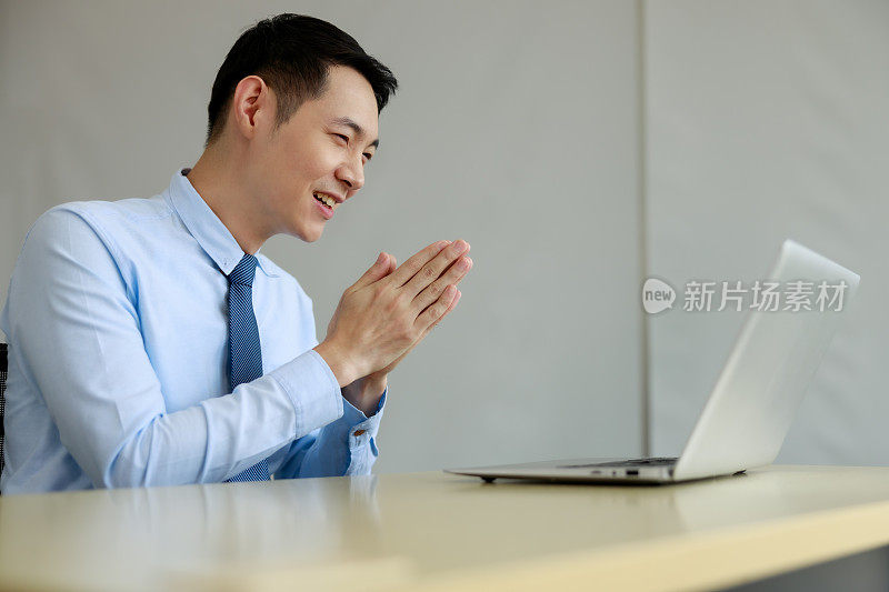 年轻的亚洲商人坐在微笑的视频电话或facetime在桌子上，举起你的手说你好问候办公室工作人员