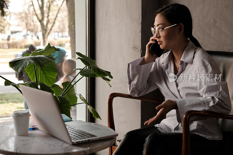 穿着衬衫的亚洲女商人在咖啡馆用手机喝茶或喝咖啡，在打开笔记本电脑前打电话。