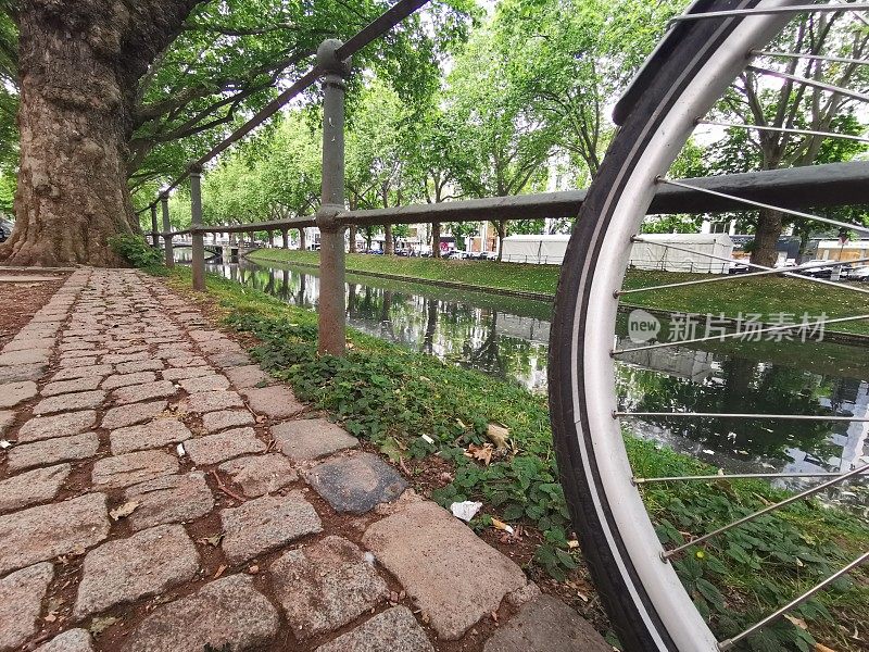 一辆自行车前轮的辐条，背景是城市护城河Königsallee，网址Düsseldorf