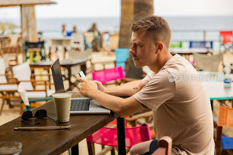 在海滩咖啡店里，男人一边用笔记本电脑工作，一边用手机