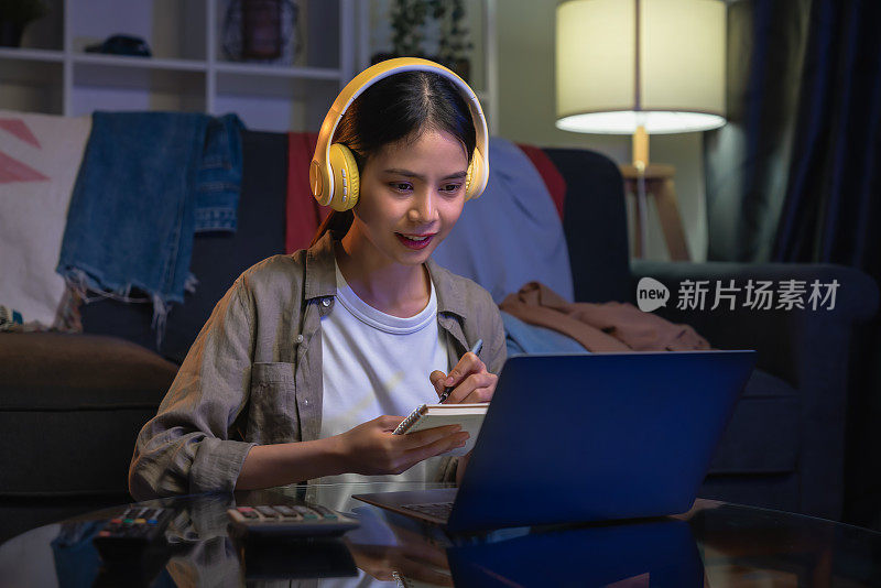 开朗美丽的亚洲女人穿着白色t恤，戴着耳机，使用笔记本电脑在视频电话会议和网上学习。