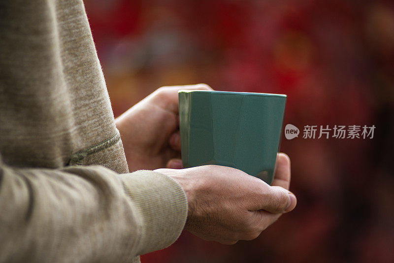 手握绿色马克杯秋茶心情红oliage模糊散景背景户外晨饮秋天舒适概念，背光度假毯。