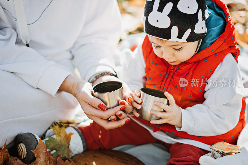 幸福的家庭母亲和蹒跚学步的小女儿在秋天野餐，用红色的保温瓶和杯子