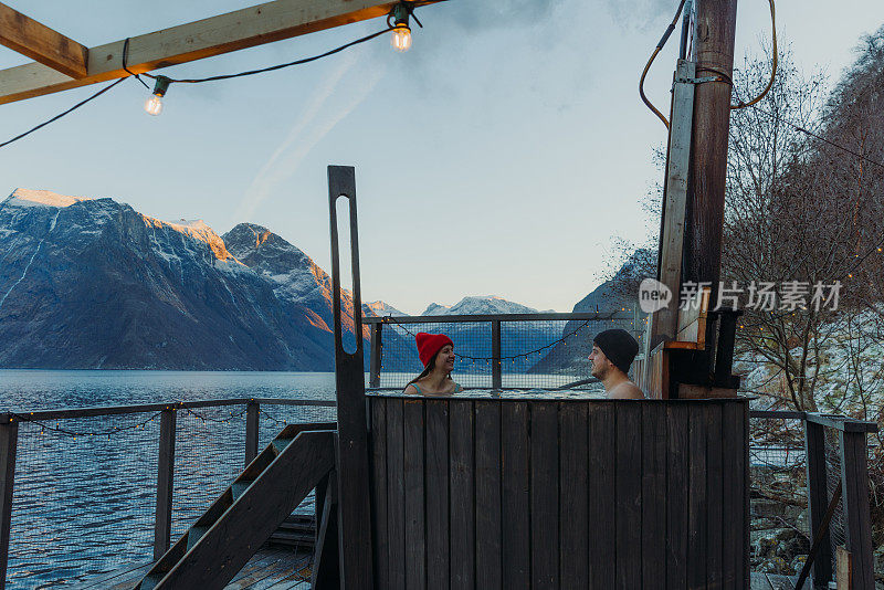 在挪威的冬天，一名女子和一名男子在峡湾边的热水浴缸里放松，欣赏风景优美的山景