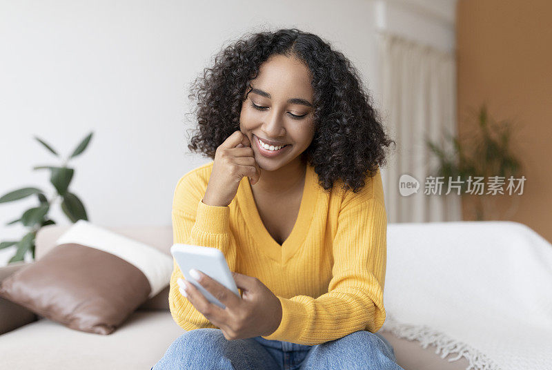 年轻的黑人女性使用手机，进行在线视频通话，坐在沙发上，在家里的社交网络上交流