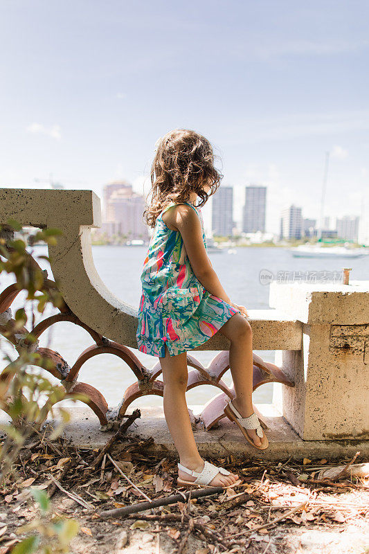 一个可爱的4岁女孩，棕色的卷发和棕色的眼睛，穿着彩色的连衣裙，帆船，白色凉鞋和白色的头发蝴蝶结，在2023年春天在佛罗里达州的棕榈滩享受探索时光