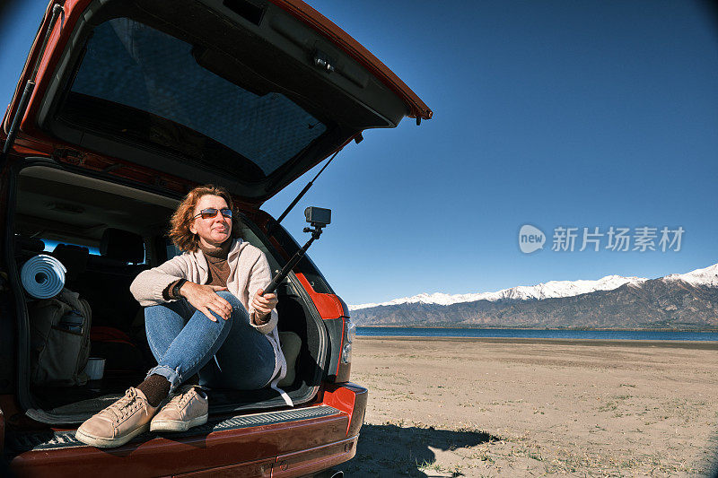 女游客坐在山湖岸的汽车引擎盖上，用运动相机拍照