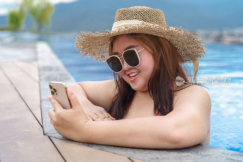 超重的年轻女子橙色swimsuitÂ在游泳池里放松地拿着智能手机。肥胖女人度假旅行用手使用手机