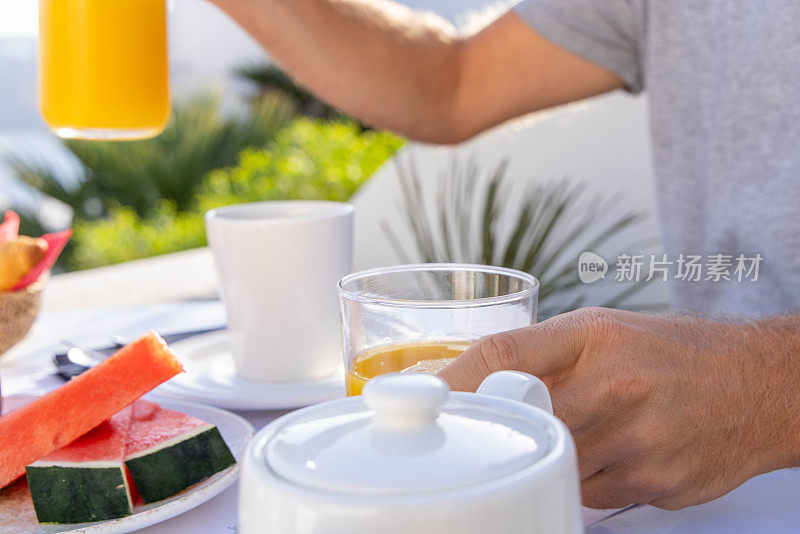 《清晨柑橘交响曲:用手将新鲜橙汁倒在以希腊风景为背景的早餐桌上》