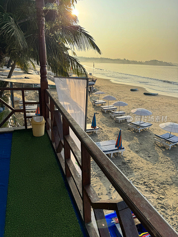 印度果阿，Palolem海滩，沙滩上一排排成对的蓝色软垫太阳躺床，遮阳伞，从海滩小屋看，人造草坪地板，露台阳台，木栏杆，重点在前景