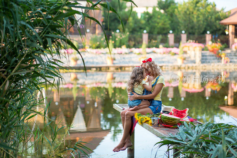 金发碧眼的母亲和三岁的女儿穿着牛仔短裤，带着红色的蝴蝶结，坐在码头上拥抱在一起，码头上有一个切好的西瓜和一束向日葵，背景是河流