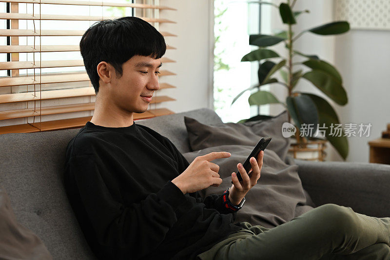 千禧一代亚洲年轻人舒舒服服坐在家里的沙发上玩智能手机的照片。