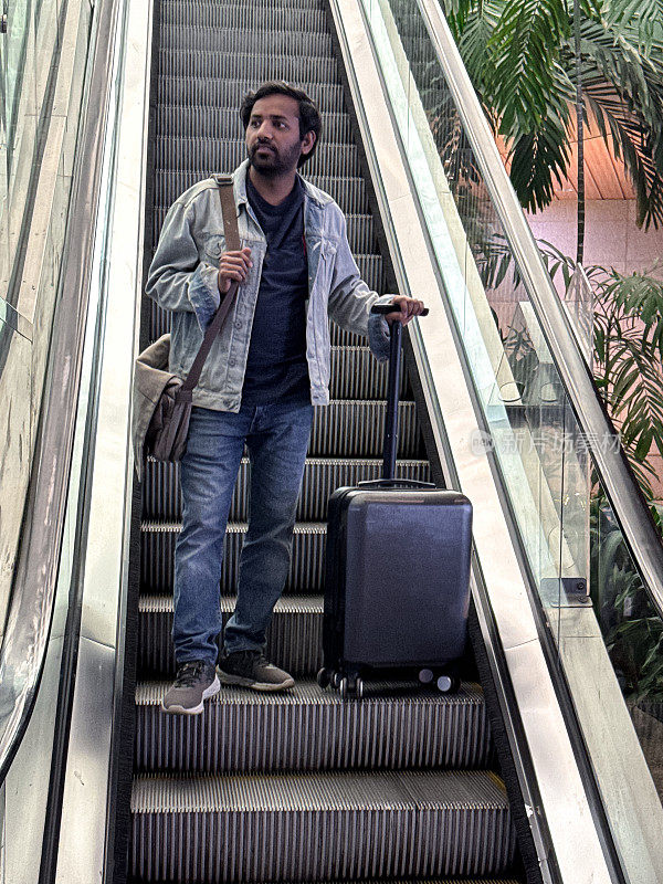 特写镜头拍摄的是印度男性旅客背着肩包在自动扶梯上旅行，带伸缩把手的轮式行李箱，机场航站楼，站在移动楼梯的金属台阶上，聚焦于前景