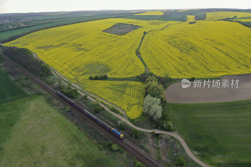 黄色油菜籽和麦田盛开，一条铁路带着蓝色列车在阴天恶劣天气下，无人机从上空鸟瞰。为石油工业提供绿色植物能源。欧洲匈牙利