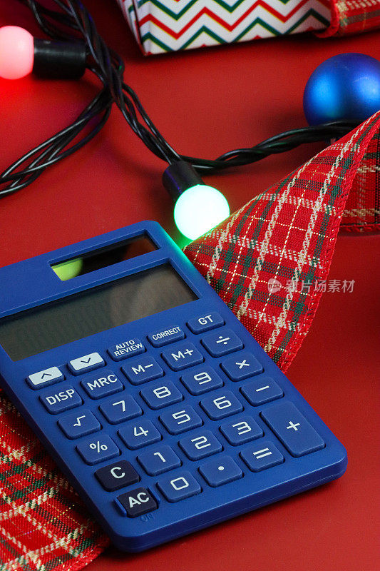 在格子丝带上的蓝色计算器的特写图像，除了照明的串在红色表面上的仙女灯，包装圣诞礼物与蝴蝶结的背景，集中在前景，圣诞节预算，账单，债务和财务概念