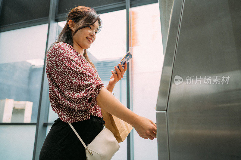 年轻的亚洲女性手持智能手机，使用自动自助邮政终端机或储物柜支付，将包裹寄存。