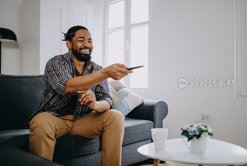 放松的年轻黑人在家里看电视，用遥控器换频道。