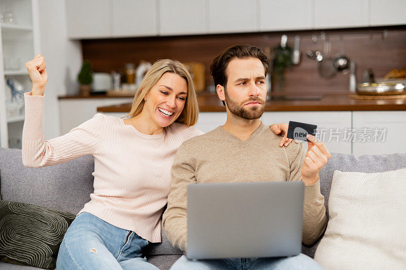 大胡子男人对花钱很不满，把钱花在网上购物上，手里拿着信用卡坐在兴奋的金发女人旁边。女孩去买东西