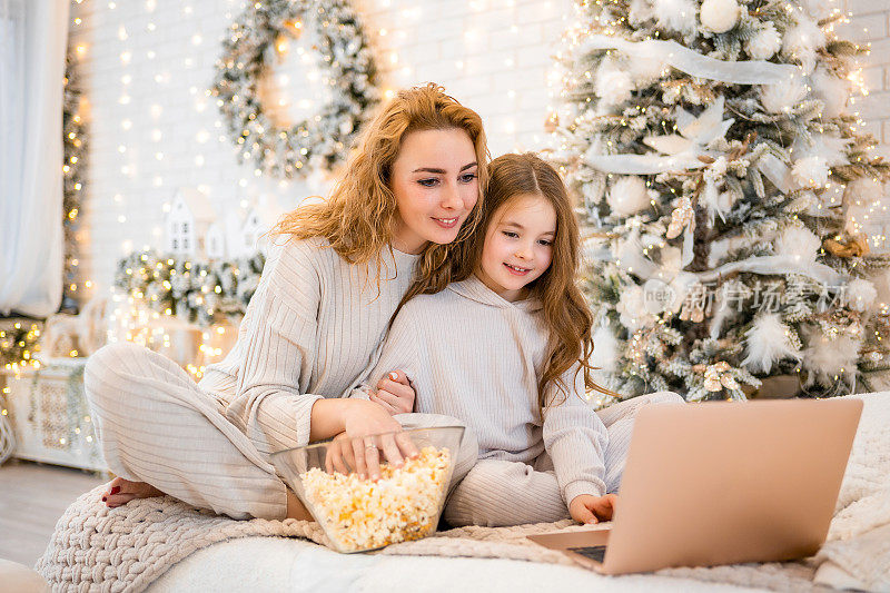 年轻女子带着7岁的小女孩穿着睡衣在圣诞装饰的家里看电影和吃爆米花