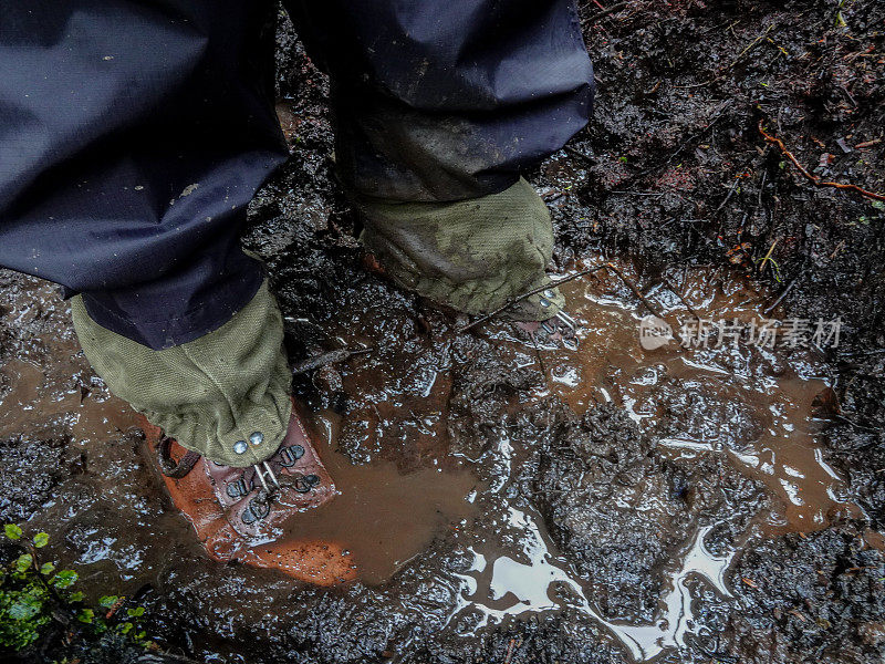 徒步靴和绑腿紧挨在泥泞的水坑里。拉基拉赛道，斯图尔特岛，新西兰。