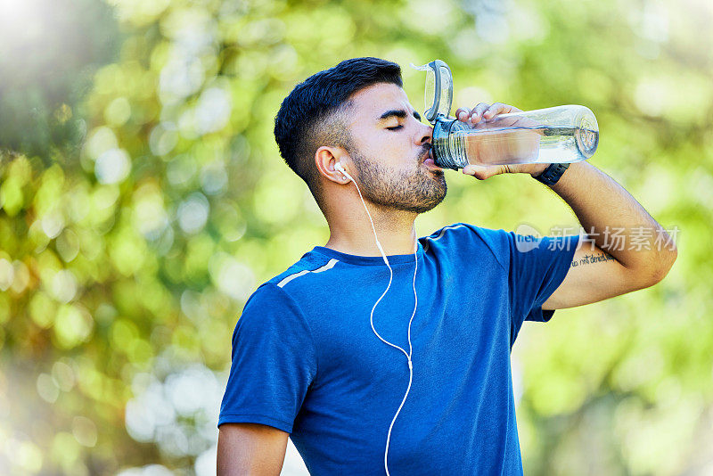 自然，跑步和口渴，一名男子在公园里戴着耳机在健身休息时喝水。健康、运动和自由，在森林中跑步，在自然景观中跑步。