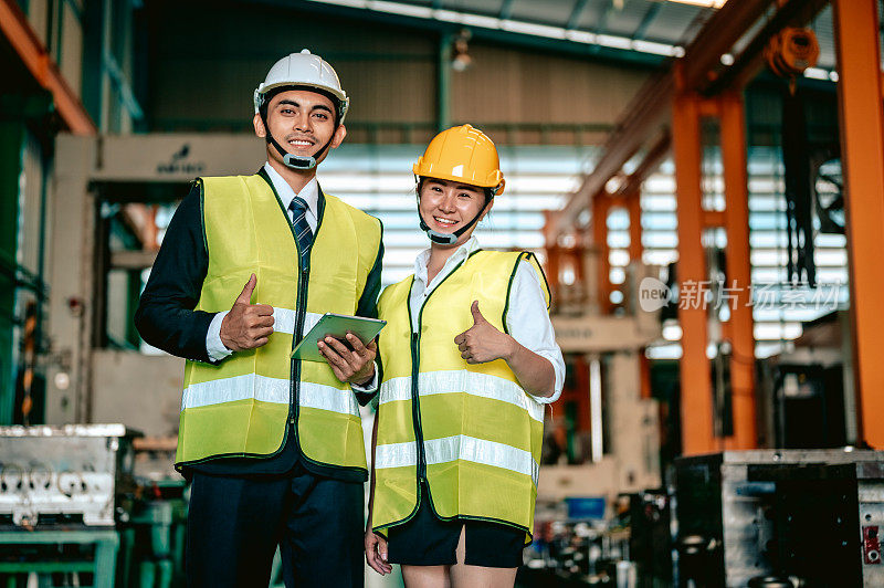 两名带着安全帽的亚洲工人在工业车间里摆姿势，微笑着竖起大拇指，看着镜头。制造业。制造业团队合作理念。
