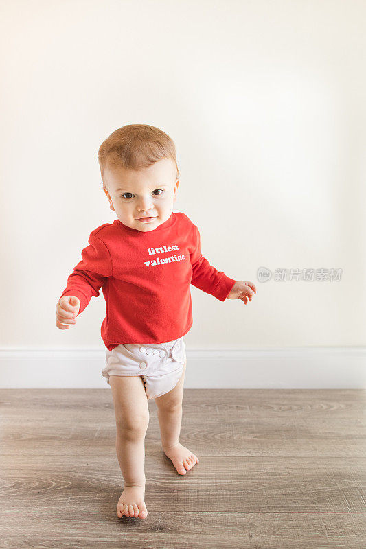 2023年2月的情人节，一个14个月大的可爱男婴赤脚站在木制硬木地板上，穿着一件写着“最小的情人节”的红色毛衣和裸色的布尿布