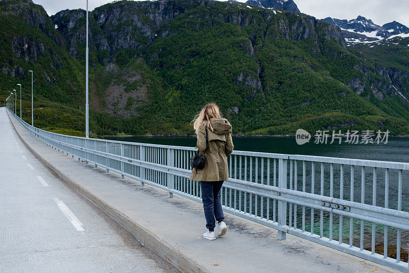 女孩沿着桥走着，欣赏着如画的风景。湖和山。挪威的旅游胜地。令人惊叹的户外风景。旅行，冒险，轻松的生活方式。罗弗敦群岛