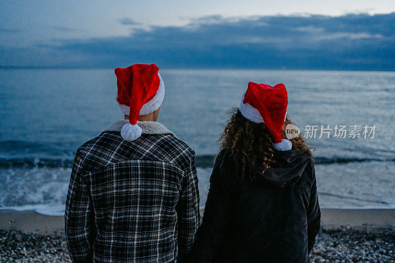 希腊，一对戴着圣诞帽的年轻夫妇在黎明时分欣赏着海岸线的景色