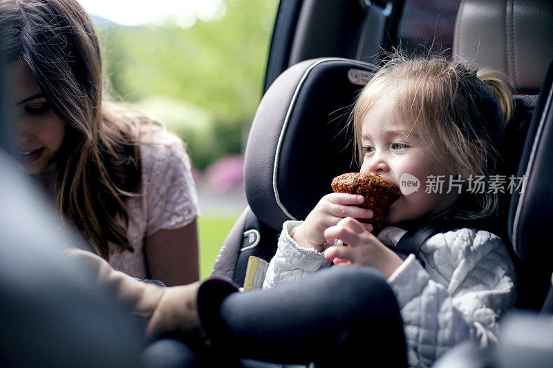 女儿吃着松饼，妈妈把安全带系在汽车座椅上