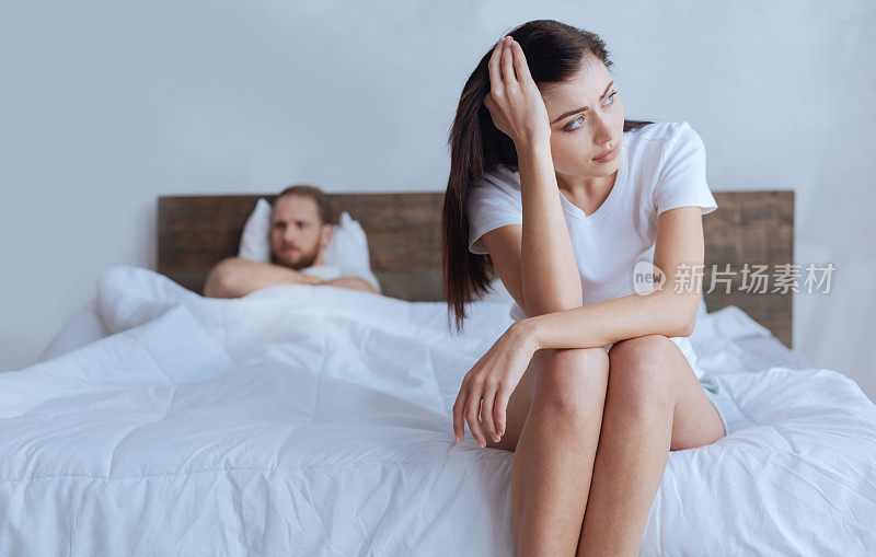心烦意乱的女人坐在床上，身后是愤怒的丈夫