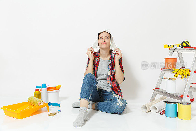 快乐有趣的女人戴着报纸帽坐在地板上，用仪器装修公寓房间隔离在白色的背景上。墙纸，胶水配件，油漆工具。修理家里的概念。
