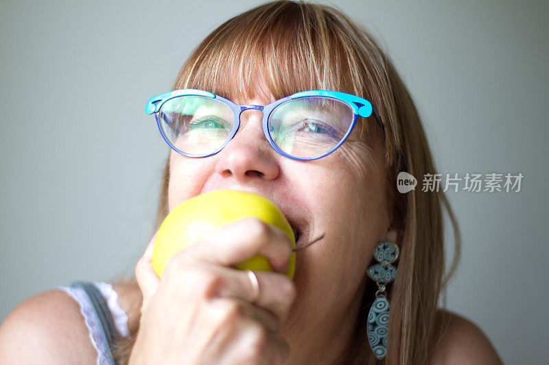 戴绿松石眼镜的快乐女人咬着健康的黄色苹果
