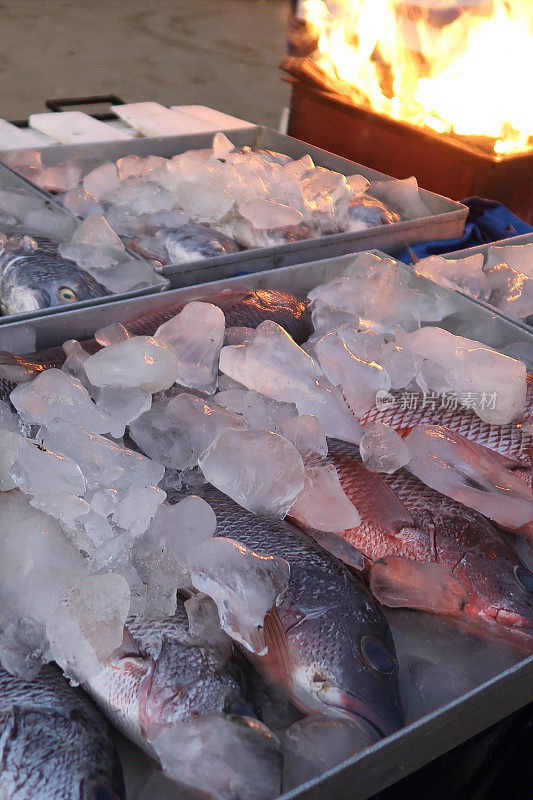 整条鱼在金属托盘的碎冰上保持新鲜，户外海滩餐厅，燃烧的烧烤