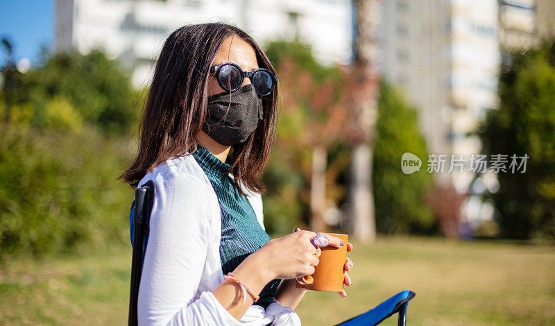 当所有的咖啡馆都因疫情而关闭时，花时间在公园喝咖啡的年轻女性遵循社交距离和口罩规则。