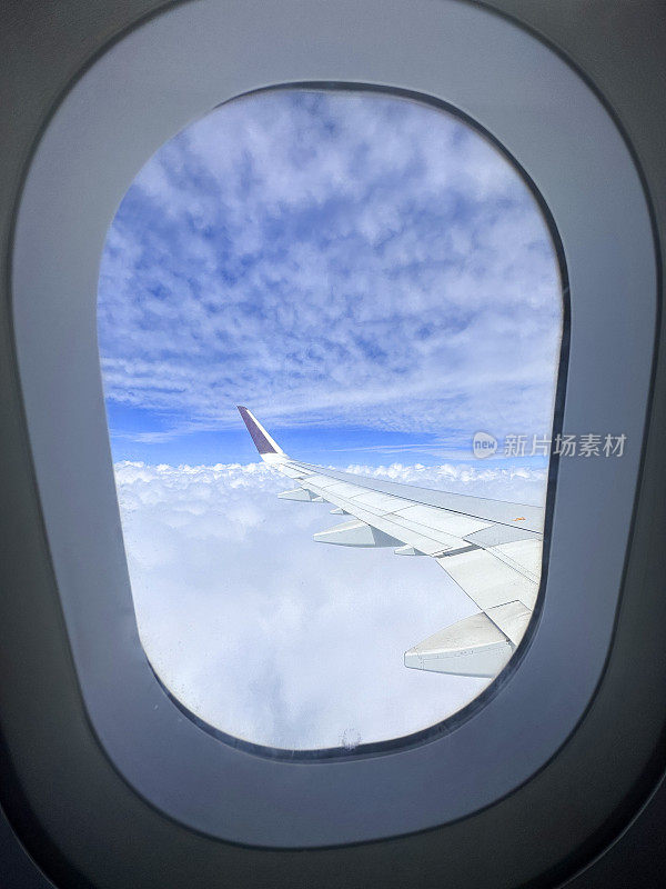 飞机舱内的窗户图像，透过飞机机翼的窗户看到云层上方，飞过蓬松的白云，小翼和板条，蓝天，聚焦于前景