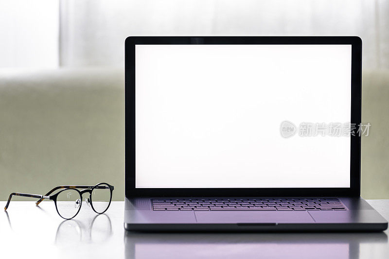 戴着眼镜的白色桌子上有一台空白屏幕的笔记本电脑。