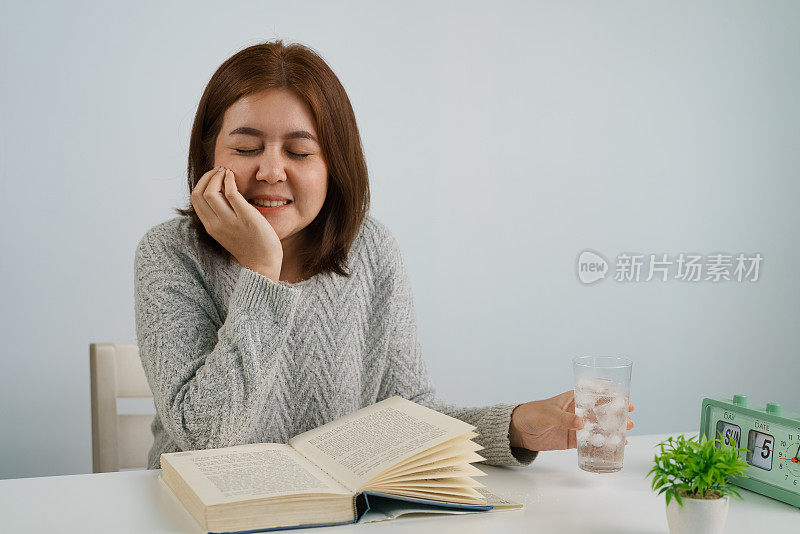 年轻的亚洲女子喝加冰的水，摸自己的脸颊，忍受牙痛，对寒冷有情绪反应。