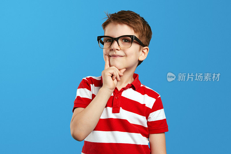 聪明的男孩戴着书呆子眼镜，穿着红色条纹衬衫，摸着脸颊，边看镜头边思考