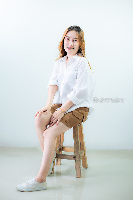 年轻的亚洲女人独自坐在白色背景的椅子上。工作室剪出的美丽短发亚洲女模特的肖像，穿着长袖白衬衫和休闲鞋，盘腿坐着。