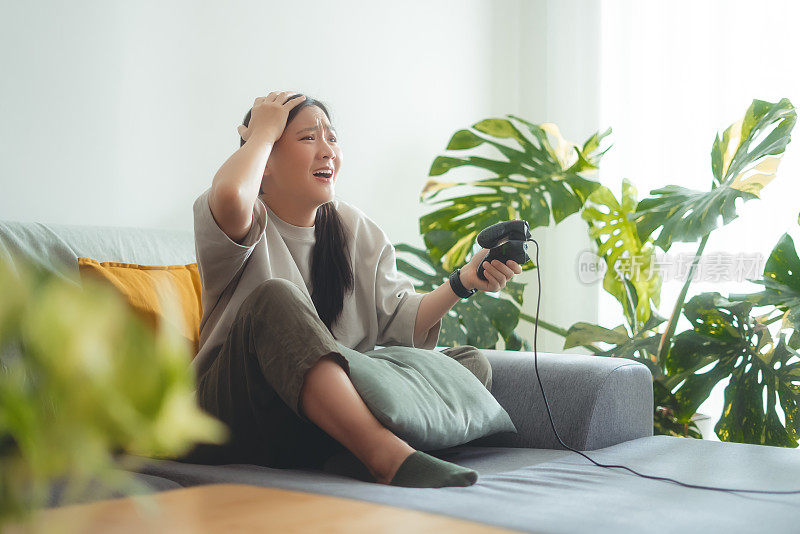 一名亚洲女子在输了游戏后坐在家里的沙发上，用操纵杆玩游戏，感到很沮丧。