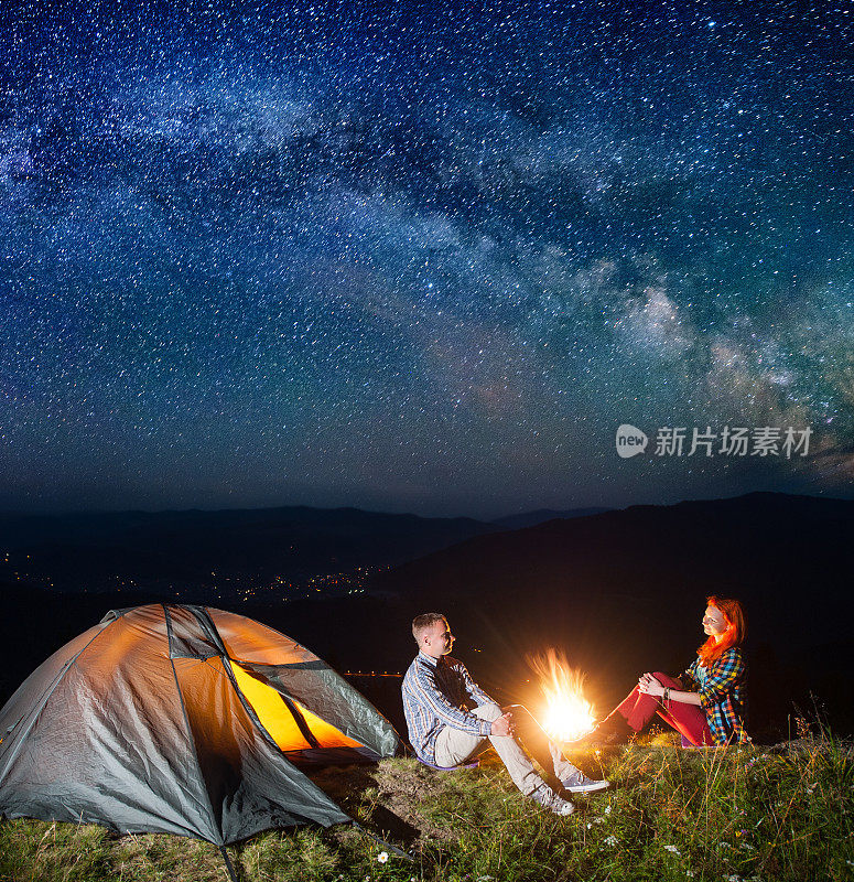 晚上露营。在令人难以置信的星空下，年轻的游客夫妇坐在篝火和帐篷旁。夜光村在山谷的背景中。天体摄影术