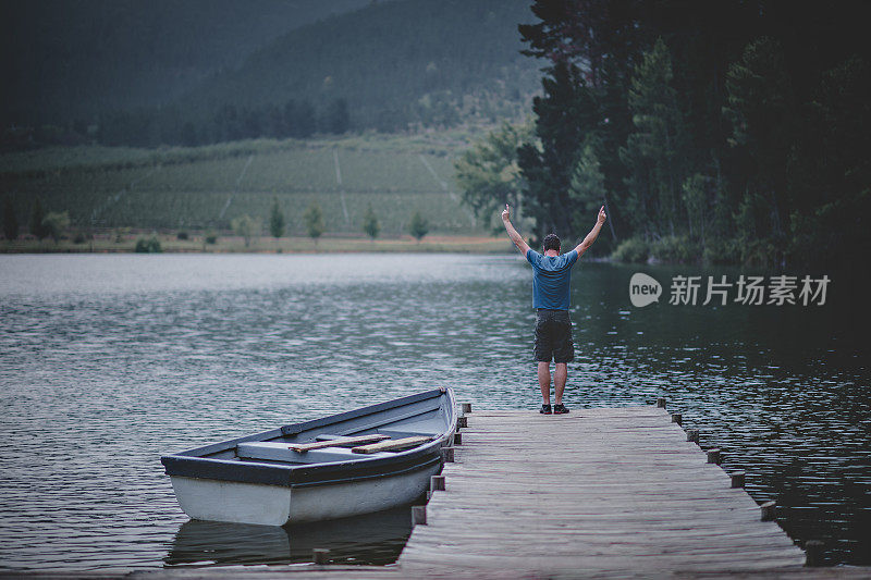 一个人举起双臂，张开双臂，站在一个美丽的湖上的木制码头上，在大自然中有一艘系泊的划艇