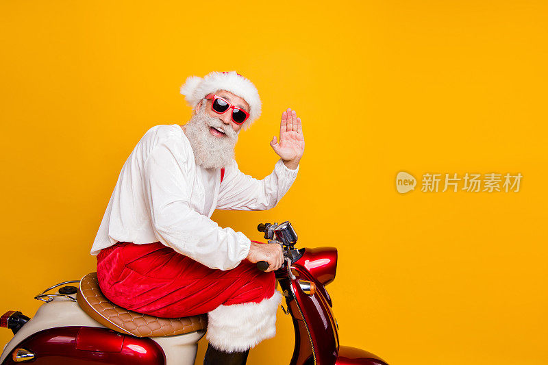 时尚的圣诞老人爷爷骑着圣诞派对复古自行车挥动手臂问候手势穿着时髦的太阳眼镜红色裤子帽子衬衫靴子孤立的黄色背景