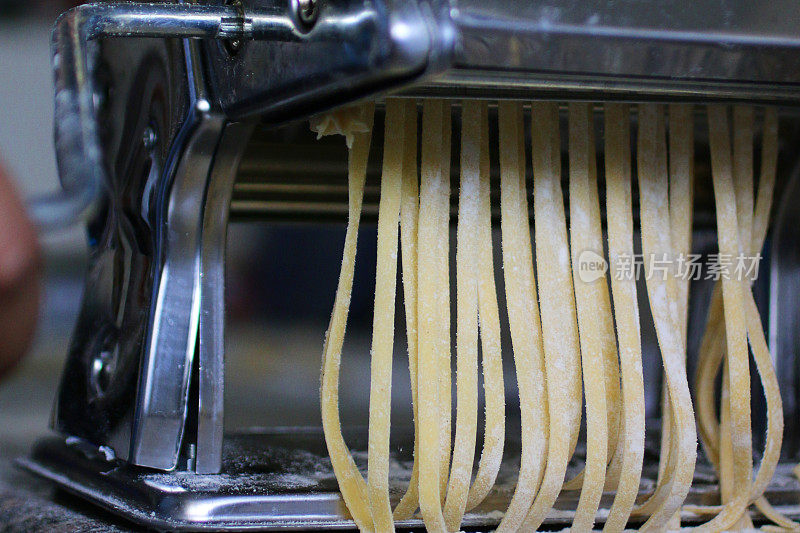 近距离拍摄的意大利面食制作机上挂着生面食的粉状带子，用擀好的面皮做家常宽面，厨房场景