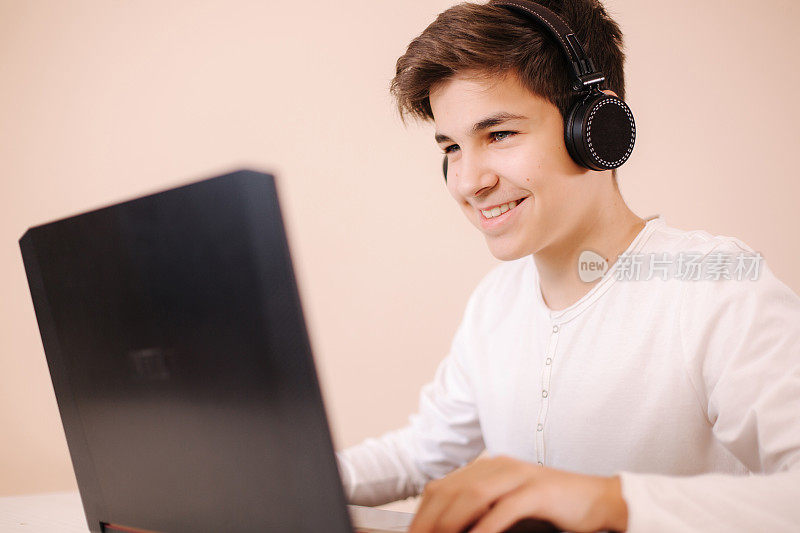 一个十几岁的男孩在房间里用笔记本电脑玩游戏。轮船使用无线耳机。快乐的玩家整天玩