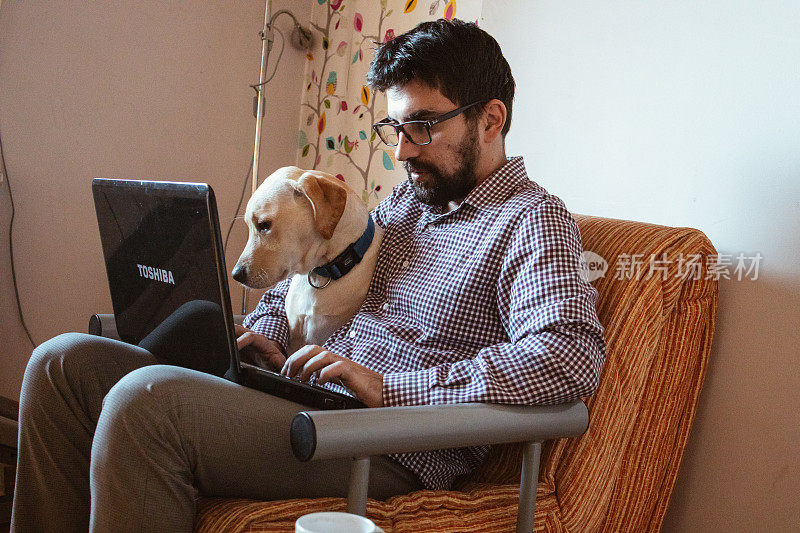 一个人在家里用笔记本电脑工作，他的宠物狗就在他旁边