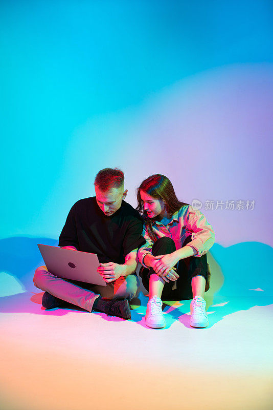 现代年轻夫妇带着笔记本电脑坐在彩色霓虹灯工作室的地板上工作、玩或网上购物