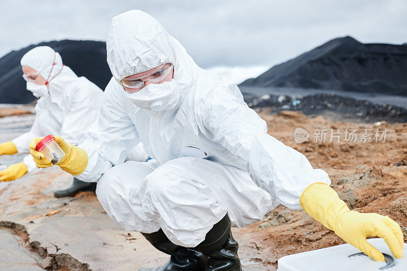 戴着呼吸器和安全护目镜的生态学家蹲在泥土地上，手里拿着泥土样品容器，在有毒区域工作